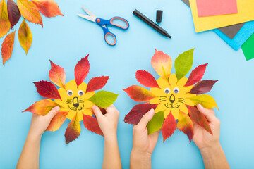Obraz na płótnie Canvas Mother and baby hands creating lion head shapes from colorful leaves and paper. Light blue table background. Making autumn decorations. Closeup. Playing and spending time together. Top down view.