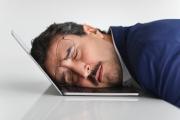 An overworked businessman in a blue suit takes a nap on his laptop keyboard in an office
