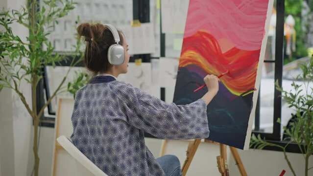 Asian woman artist listen music with headphones while painting picture on canvas creating artwork,holding tube of oil paint,fine art artist drawing at art studio.asian female painting brush on canvas