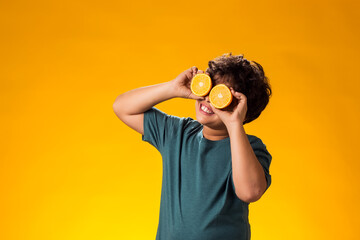 Child boy holding half of orange fruits in front of eyes over yellow background. Healthy food concept