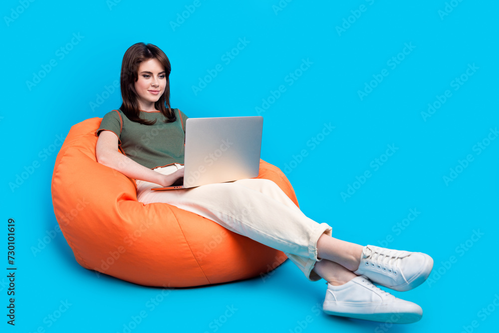 Poster full size photo of focused woman wear green t-shirt sit on pouf look at laptop chatting at distance 