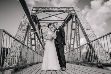 Valmiera, Latvia - July 7, 2023 - A smiling bride and groom stand close on a rusted metal bridge,...