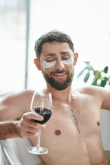 cheerful good looking man with beard and eye patches relaxing in bathtub with glass of red wine