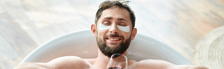 jolly attractive man with beard and eye patches relaxing in bathtub with glass of red wine, banner