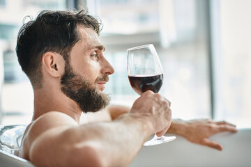 joyous handsome man with beard relaxing in bathtub with glass of red wine, mental health awareness
