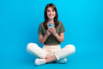 Full size photo of cheerful woman wear green t-shirt sit on floor hold smartphone read notification isolated on blue color background