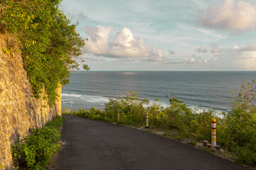 A picturesque curved road flanked by lush greenery leads to a tranquil ocean view. Safety poles...