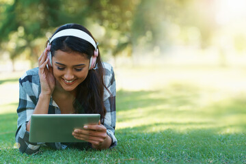 Woman, headphones and streaming music on grass, radio and internet for podcast on tablet in outdoors. Female person, happy and relaxing on lawn, weekend playlist and subscription to movie or series