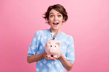 Fototapeta na wymiar Portrait of astonished positive girl open mouth arms hold money bank pig dollar bills isolated on pink color background