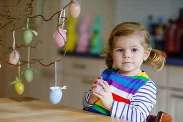 Cute little toddler girl decorating tree bough with colored pastel plastic eggs. Happy baby child having fun with Easter decorations. Adorable healthy smiling kid in enjoying family holiday