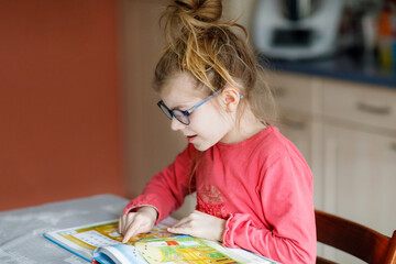 Little girl with eyeglasses learning reading. Child of elementary school reads a book at home,...