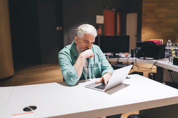 Pensive senior Caucasian man in green shirt working on laptop in contemporary office environment, reflecting concentration, problem-solving, and concept of continuous professional development.