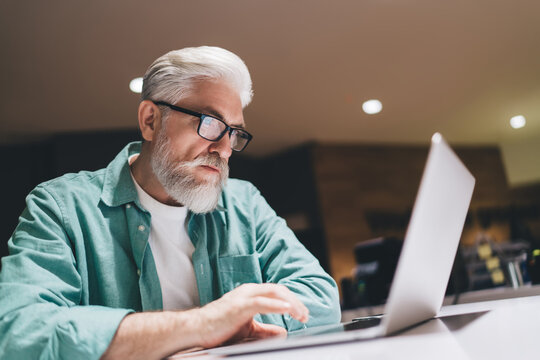 Focused senior Caucasian man with a beard working intently on a laptop in an indoor setting, wearing glasses and a green shirt, symbolizing professional dedication and digital engagement in later life