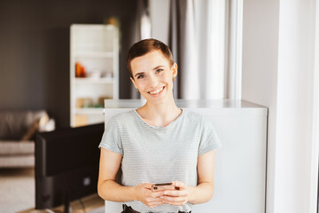 Young woman with short hair laughing while holding smartphone in her apartment - 730178682