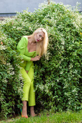 Full-length portrait of a beautiful young blonde with long hair in a yellow-green pantsuit and bright high-heeled pumps against a background of green bushes with flowers