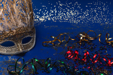 Golden Mardi Gras or carnival face mask on a dark blue background with colored tinsel and confetti....