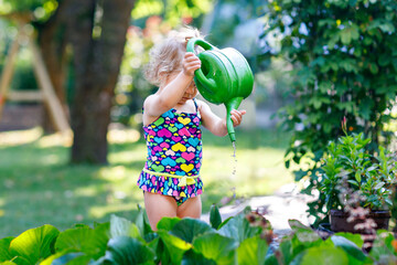 Cute little baby girl in colorful swimsuit watering plants and blossoming flowers in domestic...