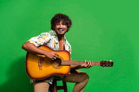 Photo portrait of attractive young man sit chair play guitar dressed stylish citrus print clothes isolated on green color background - Powered by Adobe