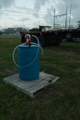 Vertical blue 55 GALLON PLASTIC DRUM ON old weathered pallet  WITH HAND WATER PUMP. In a grass field with electric substation and utility vehicles in the background. Has a blue sky with white clouds.
