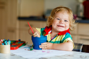 Cute adorable baby girl learning painting with water colors. Little toddler child drawing at home,...