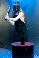 A girl in a protective mask smashes a glass bottle with a baseball bat in the destruction room to...