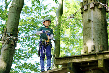 Naklejka na ściany i meble Two children in forest adventure park. Kids boys in helmet climbs on high rope trail. Agility skills and climbing outdoor amusement center for children. Outdoors activity for kid and families.