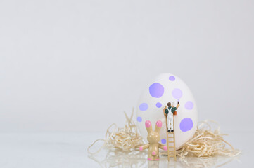 A painter on a wooden ladder paints a white egg for Easter, an Easter bunny looks on, white...