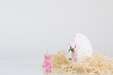 A painter on a wooden ladder paints a white egg for Easter, an Easter bunny looks on, white background
