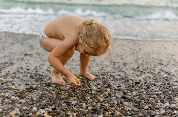 A child collecting wet pebbles on the seashore. A barefoot kid girl playing with stones pebbles near water waves. Collecting stones, intellectual curiosity.