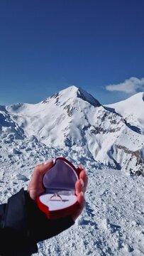 Female hand closeup holding an engagement ring inside a red heart shaped box over the snowy mountain peaks background. Marriage proposal on the mount top