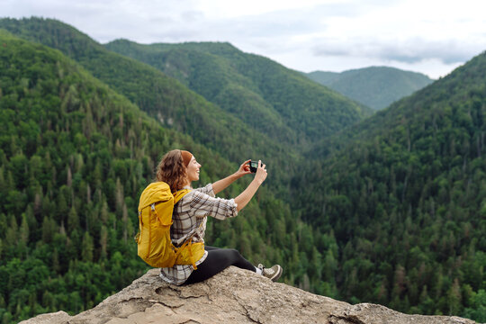 Tourist woman taking picture outdoors for memories, making selfie on top of cliff with valley mountains view, sharing travel adventure journey. 