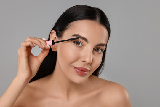 Beautiful young woman applying mascara on grey background