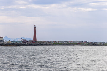 A towering red lighthouse, a beacon of Andenes, Norway, rises above a calm sea with a backdrop of a cozy, colorful town.
