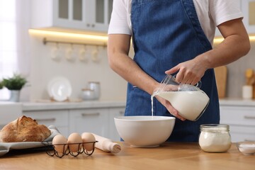Making bread. Man pouring milk into bowl at wooden table in kitchen, closeup