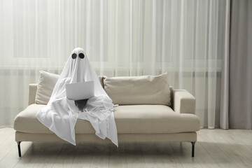 Creepy ghost. Person covered with white sheet using laptop on sofa at home, space for text