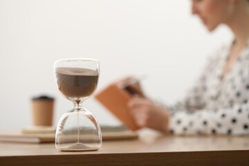 Hourglass with flowing sand on desk. Woman working indoors, selective focus