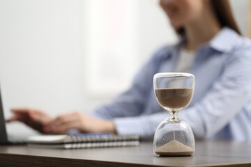 Hourglass with flowing sand on desk. Woman using laptop indoors, selective focus