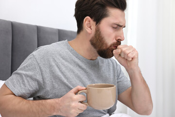 Sick man with cup of hot drink coughing on bed at home. Cold symptoms