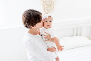 mom gently hugs her baby daughter kissing and admiring them, happy family, mom with baby on the bed, maternal love and care, mom on maternity leave playing with the baby