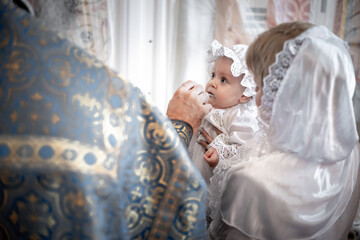 anointing of a small child in an Orthodox Christian church or temple during the sacrament of baptism with a priest or priest in the mother's arms - obrazy, fototapety, plakaty