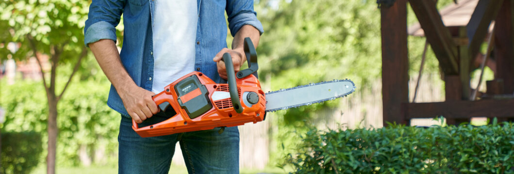 Unrecognizable craftsman standing with cordless chain saw, working in summer garden. Crop view of strong handyman in denim clothes holding modern electric saw. Concept of garden equipment. 