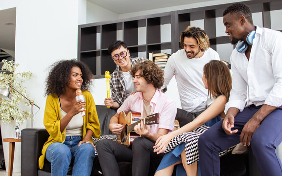 Group of diverse friends or people wearing casual clothes, relax smiling with happiness, party celebration, sitting in living room at cozy home, playing guitar and singing. Birthday, New Year Concept.