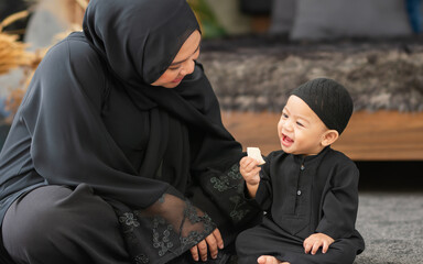 Muslim mother wearing traditional black dress and headscarf, feeding food to baby boy, eating with...