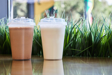 Two clear plastic glasses with lids filled with chocolate vanilla milkshakes on table, green grass....