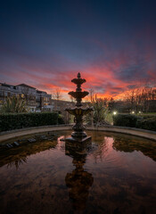 Fountain and pond in a park at sunset with a pink sky. Evening view in portrait orientation with buildings and trees behind.
