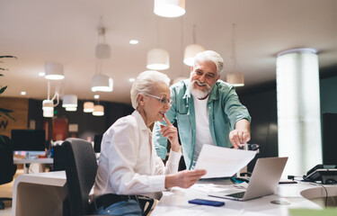 Joyful senior Caucasian male and female in 60s, review documents together over laptop in office. He is wearing a teal shirt, pointing at the paper, while she, in a white blouse, examines it closely