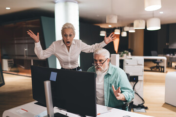Shocked senior Caucasian man and woman in office setting, reacting to computer screen. Man in teal...