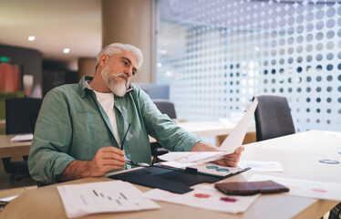 Pensive senior Caucasian man with white beard analyzing financial documents at a modern office desk, dressed in a casual teal shirt, showing a moment of contemplation