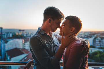Young happy cute gay couple hugging and kissing on balcony overlooking city and sunset - obrazy, fototapety, plakaty