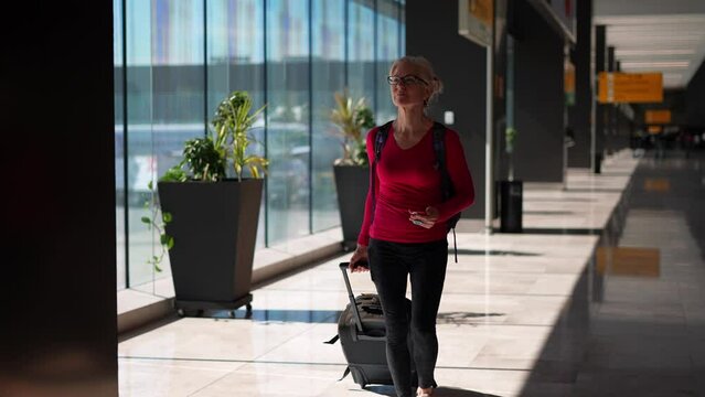 Happy attractive blond woman wearing backpack pulling luggage holding phone walking towards camera in an airport.